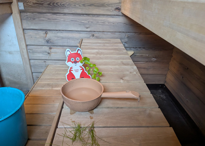 Inne i en bastu. En räv i papper är omringad av grönt gräs och blad.